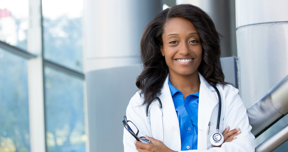 Smiling female graduate nurse in white coat with stethoscope standing in hospital